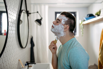 Young man with down syndrome learning how to shave, applying shaving foam all over his face,...