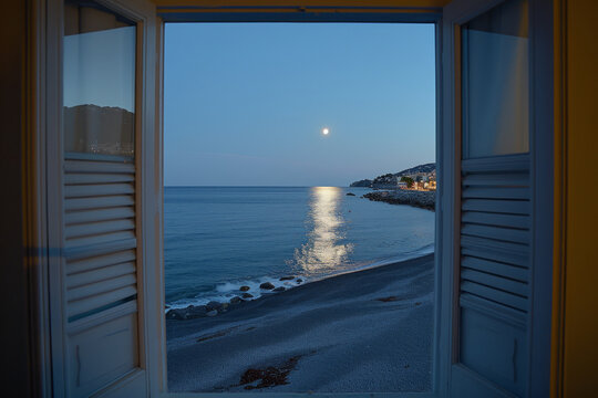 Fototapeta View through an open window with shutters, to see a sandy beach, rocky coastline, lovely small town and beautiful blue sky with big, silver fully moon at blue hour...