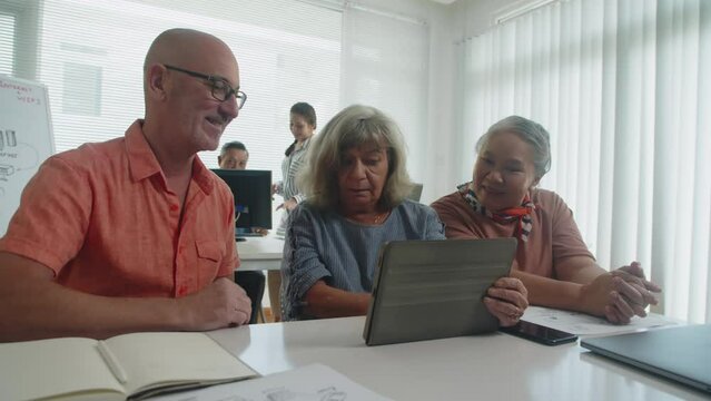 Medium shot of mature woman explaining to same age students digital tablet features during group lesson