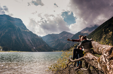 Young couple taking selfie through smart phone on fallen tree trunk near lake Vilsalpsee and mountains at Tyrol, Austria