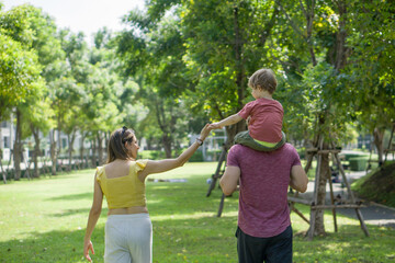 Naklejka na ściany i meble A woman, her husband and her son walk together, enjoying nature in a sunny park with trees and a walking path. View from behind