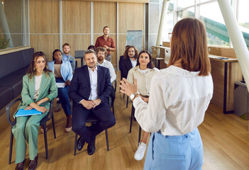 Confident female business coach teaches small group of people new business knowledge. People are sitting on chairs in office of business center listening to woman standing with her back to camera. - obrazy, fototapety, plakaty