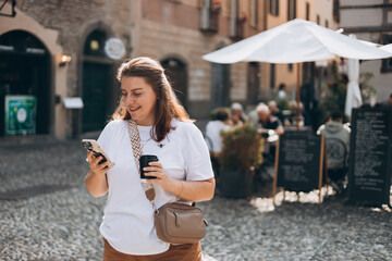 Happy young Caucasian woman in good mood walks with coffee and phone around city during day....