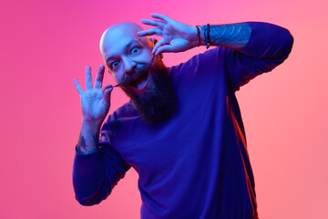 Gel portrait of bearded bald man in blue sweater expressing emotions, grimacing against pink background in neon light. Concept of human emotions, facial expression