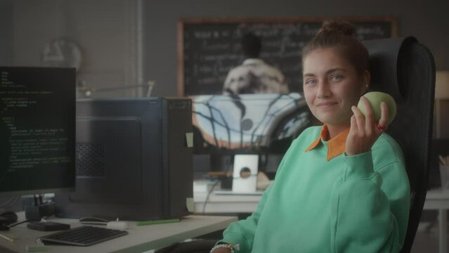 Portrait of young female software tester holding green apple and smiling at camera while sitting at desk with computer during office workday