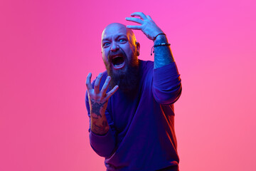 Portrait of emotional breaded bald man expressing fearful face, gesturing, shouting against pink background in neon light. Concept of human emotions, facial expression