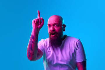 Emotive shocked beard bald man raising finger up, expressing ideas against blue background in neon...