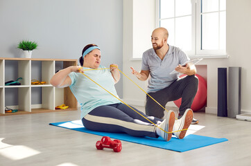 Overweight woman doing fitness workout with her trainer in fitness club. Plus size, obese, fat woman in sportswear sitting on mat training with resistance band under guidance of personal trainer