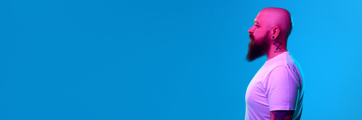 Portrait of bearded bald man with tattoo on neck, in casual white t-shirt posing against blue...