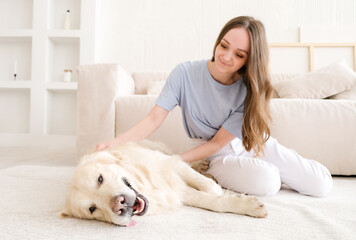 Portrait of happy young woman with pet dog sitting on floor at home. Caucasian female hugging golden retriever in living room