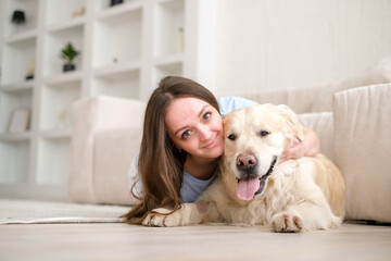 Portrait of happy young woman with pet dog, sitting on floor at home. Caucasian female hugging golden retriever in living room
