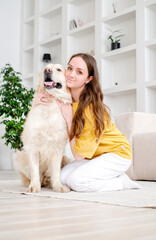 Portrait of happy young woman with pet dog, sitting on floor at home. Caucasian female hugging golden retriever in living room