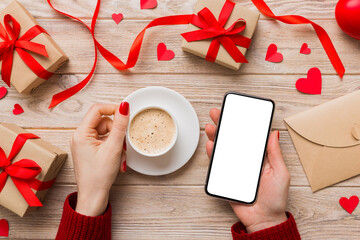 Female hands holding smart phone with coffee for Valentine day, gift box and envelope, hearts