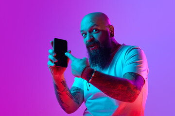 Online services. Bearded bald man with tattoo, pointing on mobile phone against gradient purple...
