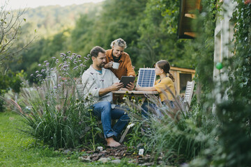 Parents with daughter sitting outdoors in garden, with model of solar panel, learning about solar...