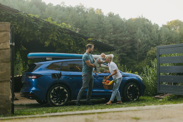Young family with baby unloading groceries from their electric car. Electric vehicle with charger...