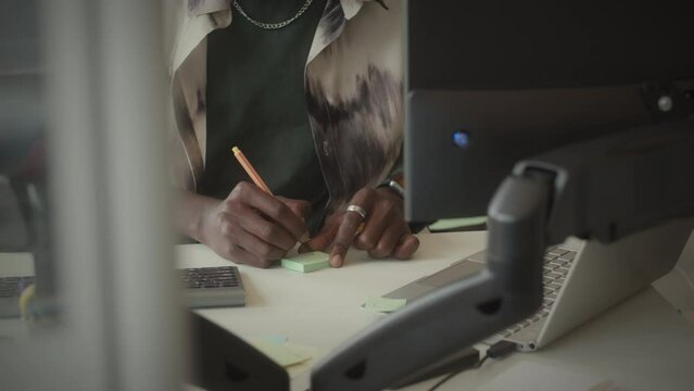 Tilt up shot of young Black male programmer with neck pillow writing on sticky note, attaching it to monitor and working on computer in office