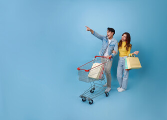 Full size young cheerful Asian couple have fun drive shopping trolley cart and holding shopping bag shopping grocery in mall isolated on blue background © NaMong Productions
