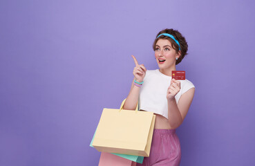 young attractive shopper woman carrying shopping bag and credit card with hand pointing finger to copy space isolated on purple background.