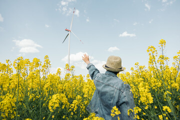 Teenage girl with her back turned with her hand up pointing at a wind turbine in a field of yellow...