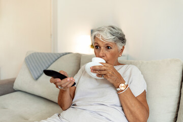 Senior woman, sofa and coffee to relax with remote for television, video or movies with smile in...