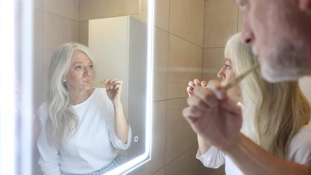 couple reflected in the mirror while brushing their teeth, the focus is on the older woman with gray hair