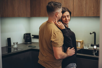 A young couple is standing in the kitchen. The guy sneaks up behind the girl and touches her waist with his hand. The girl, closing her eyes in a half-turn, touches the guy’s cheek with her fingers - obrazy, fototapety, plakaty