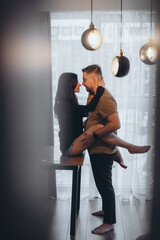 The girl sits on the table with her legs around the guy and holds him by the neck. They look at each other. Blurred background and foreground with highlights