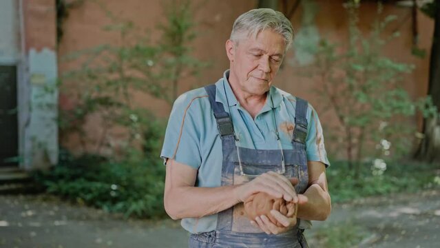 Knead grinding clay dought for pottery wheel session. Old man professional handicraft master preparing to create artwork. Elderly male potter master kneading the raw clay outdoors