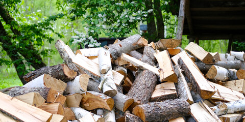 A big pile of stacked freshly chopped firewood for kindling against a background of green bushes and trees. Concept of firewood preparation, deforestation, heating. Panoramic advertising banner