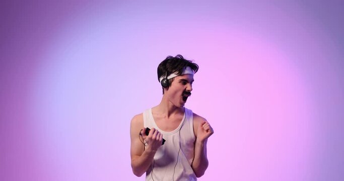 Crazy and funny Caucasian young 90s man disco dancing with retro personal stereo player, listening music using headphones on purple background.
