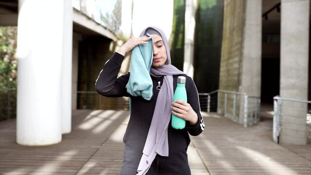 muslim woman in hijab wearing sportswear resting drinks water and dries with towel