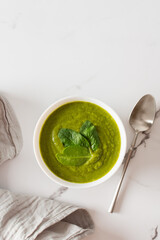 Healthy breakfasts, green broccoli and spinach cream soup in a white bowl on a marble background with a gray napkin, top view