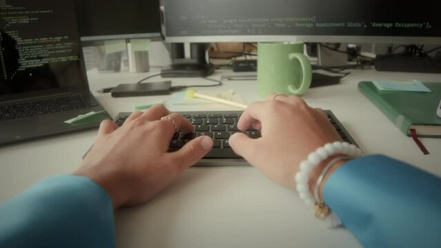 POV shot of hands of female programmer using wireless keyboard while coding on computer and drinking tea from mug at desk in office