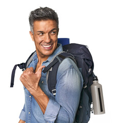 Hispanic hiker with backpack and flask points with thumb finger away, laughing and carefree.