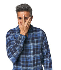 Middle-aged Latino man blink at the camera through fingers, embarrassed covering face.