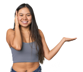 Young Chinese woman in gray top, studio holds copy space on a palm, keep hand over cheek. Amazed and delighted.