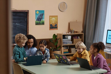 Diverse group of little children using computers at table in school classroom with female teacher...
