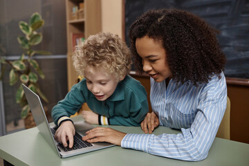Portrait of young Black female teacher with little boy using laptop computer in school classroom
