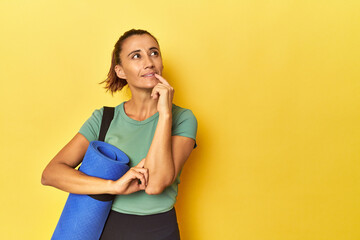 Middle aged sportswoman with yoga mat on yellow studio looking sideways with doubtful and skeptical...