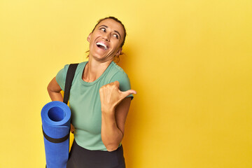 Middle aged sportswoman with yoga mat on yellow studio points with thumb finger away, laughing and...