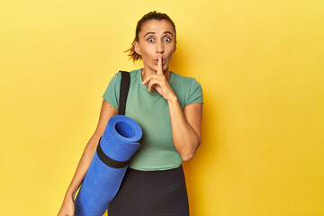Middle aged sportswoman with yoga mat on yellow studio keeping a secret or asking for silence. - obrazy, fototapety, plakaty