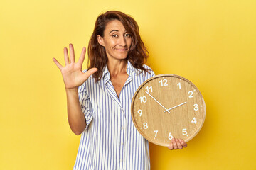 Middle aged woman holding a wall clock on a yellow backdrop smiling cheerful showing number five...