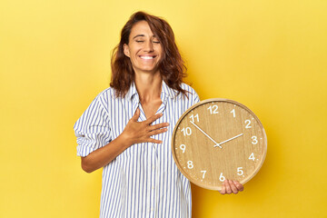 Middle aged woman holding a wall clock on a yellow backdrop laughs out loudly keeping hand on chest.