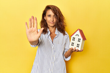 Middle aged woman holding a miniature house on yellow backdrop standing with outstretched hand...