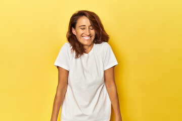 Middle-aged woman on a yellow backdrop laughs and closes eyes, feels relaxed and happy.