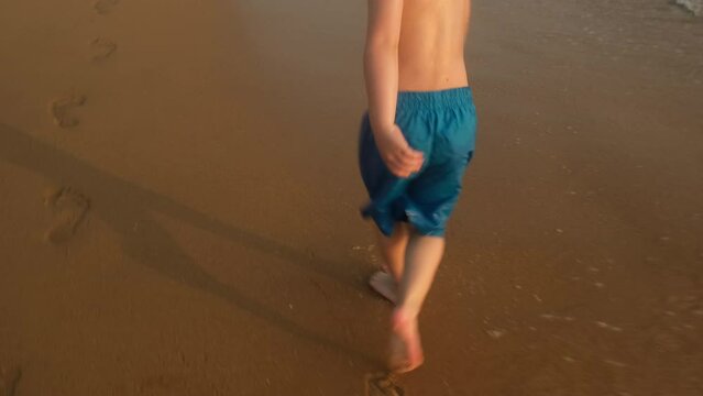 Small boy having fun running barefoot along ocean coast at sunset.