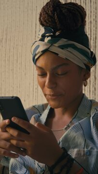 Vertical closeup of young cheerful African American lady in durag looking at phone and smiling while chatting indoors