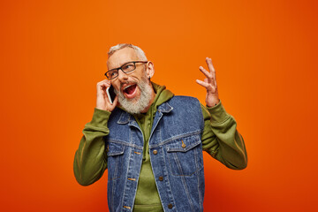 Fototapeta premium good looking joyful mature man in vibrant green hoodie and denim vest with glasses talking by phone