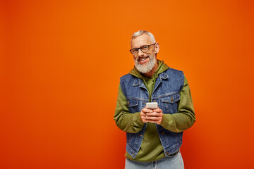 Obraz premium joyous handsome mature male model in vibrant attire holding smartphone and smiling at camera